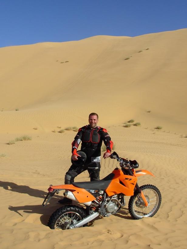 Ein Mann posiert mit seinem orangefarbenen Motorrad in einer Sandwüste.
