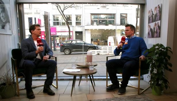 Zwei Männer sitzen in Stühlen und sprechen in Mikrofone vor einem Fenster mit Blick auf eine belebte Straße.