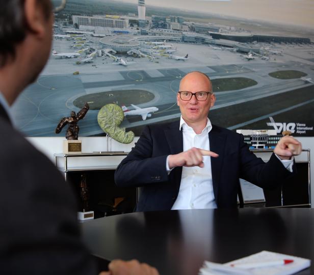 Ein Mann gestikuliert in einem Gespräch vor einem Bild des Vienna International Airport.