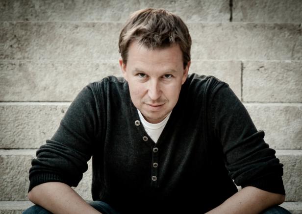 Ein Mann mit braunen Haaren sitzt auf einer Steintreppe und blickt in die Kamera.