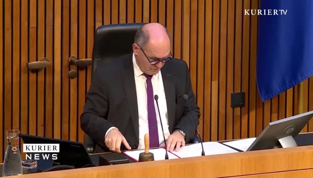 Ein Mann in einem Anzug sitzt an einem Tisch mit einer Glocke und Dokumenten.