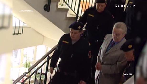 Ein Mann in einem hellen Anzug wird von uniformierten Beamten eine Treppe hinuntergeführt.