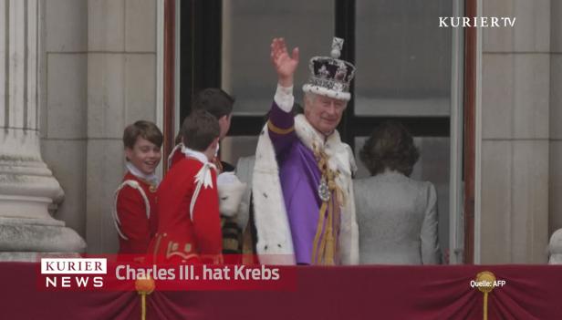 König Charles III. mit Krone und Hermelinmantel auf einem Balkon, winkend.