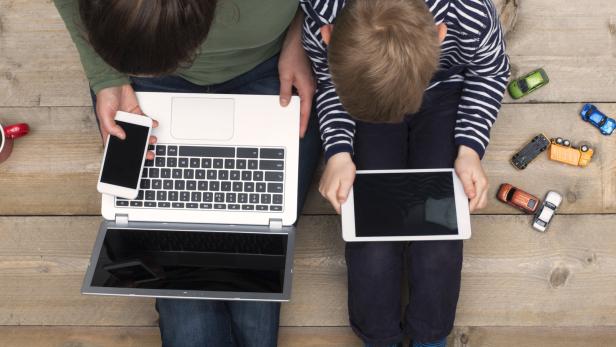 Eine Frau mit Laptop und Handy sitzt neben einem Kind mit Tablet auf einem Holzboden.