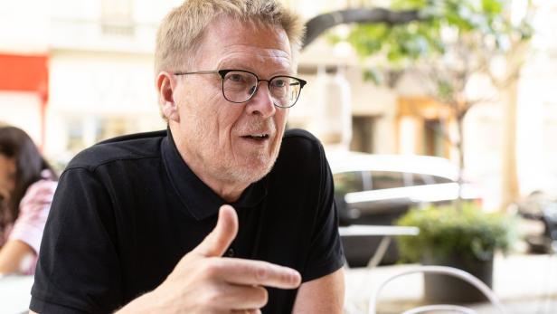 Ein Mann mit Brille gestikuliert an einem Tisch im Freien.