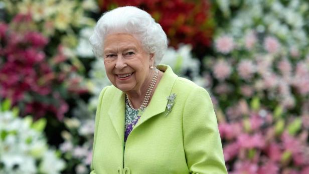 Königin Elizabeth II. lächelt in einem hellgrünen Kostüm mit Brosche vor einem Blumenmeer.