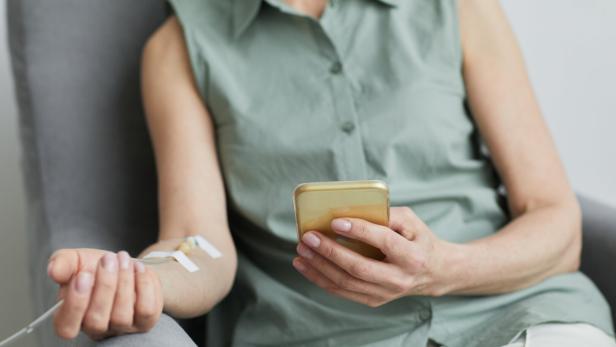 Eine Person mit Infusionskanüle im Arm hält ein Smartphone.
