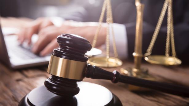 Ein Richter tippt am Laptop, im Vordergrund Hammer und Waage auf einem Holztisch.