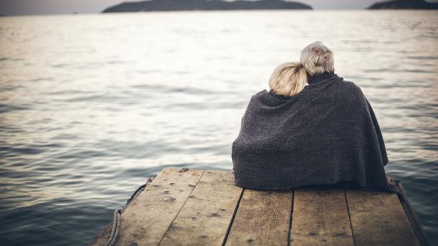 Ein älteres Paar sitzt gemeinsam unter einer Decke auf einem Holzsteg am Meer und blickt auf das Wasser.