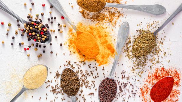 Auf einer weißen Oberfläche arrangierte Löffel sind mit verschiedenen Gewürzen wie Paprika, Curry und Pfefferkörnern gefüllt.