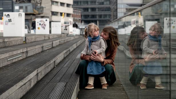 Eine Mutter umarmt ihre kleine Tochter mit Brille auf einer Treppe in einer städtischen Umgebung.