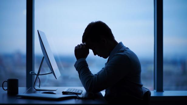 Eine Person sitzt mit gesenktem Kopf und verschränkten Händen nachdenklich vor einem Computer am Schreibtisch.