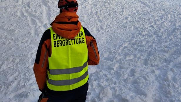Ein Mitglied der Bergrettung steht vor einem schneebedeckten Hang mit Suchmannschaft.