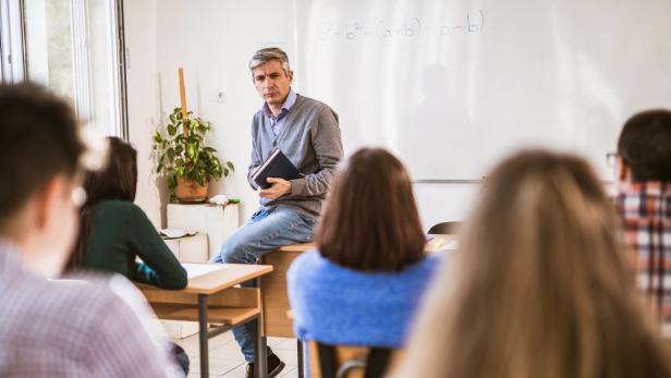 Ein Lehrer sitzt auf einem Tisch vor einer Klasse und hält ein Buch.