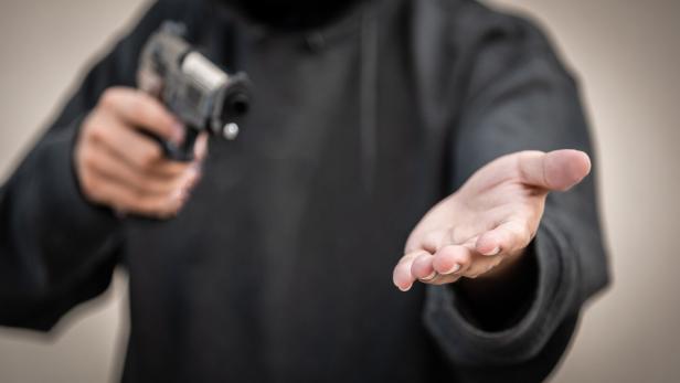 Eine Person in einem schwarzen Hoodie hält eine Pistole in der einen und eine offene Hand in der anderen.