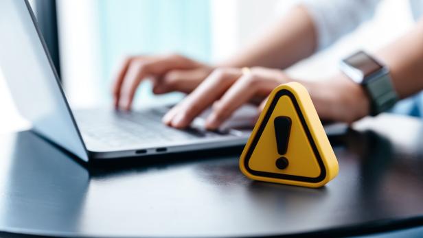Ein gelbes Warnschild mit Ausrufezeichen steht vor einem Laptop, an dem eine Person tippt.