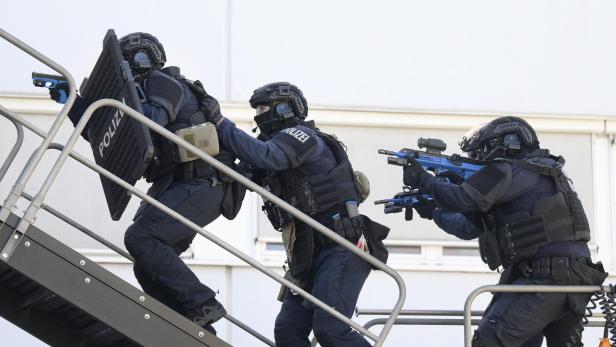 Drei Polizisten in voller Montur steigen eine Treppe hinauf, einer mit Schild, einer mit blau gefärbter Waffe.