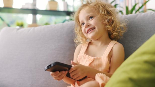 Ein lächelndes Mädchen mit blonden Locken sitzt mit einem Smartphone auf einem Sofa.