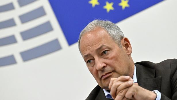 Ein Mann sitzt vor einer Europaflagge und blickt nachdenklich zur Seite.
