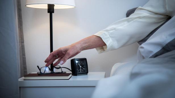 Eine Hand greift nach einer Brille auf einem Nachttisch mit Buch, Wecker und eingeschalteter Lampe.