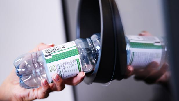 Eine Hand wirft eine leere Plastikwasserflasche in einen Recyclingbehälter.