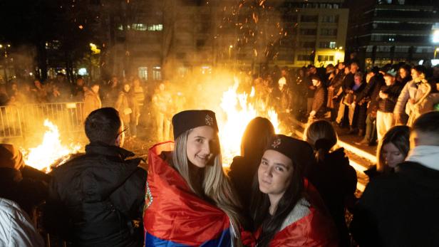 Zwei junge Frauen mit serbischen Flaggen stehen vor einem Lagerfeuer.