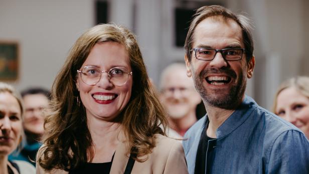 Eine lächelnde Frau mit Brille und ein lachender Mann stehen vor einer Gruppe von Menschen.
