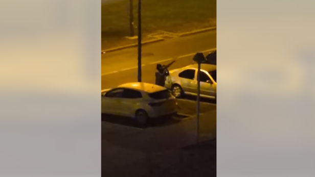 Eine Person zielt mit einer Waffe in einer dunklen Straße neben geparkten Autos.