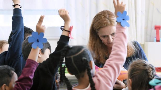 Mehrere Kinder heben die Hand, während eine Frau ihnen aufmerksam zuhört, einige Kinder tragen gebastelte Blumen am Handgelenk.