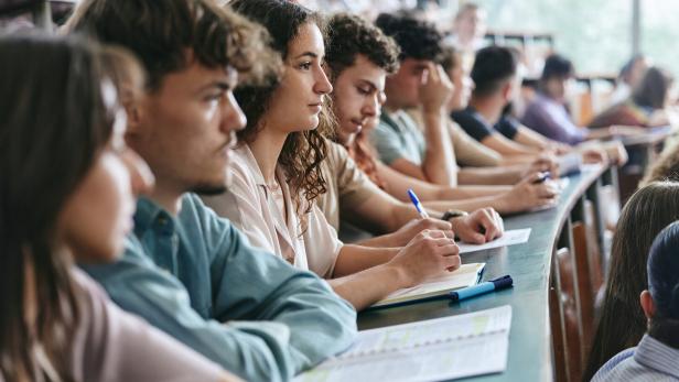 Man sieht junge Studierende in einem Vorlesungssaal. Sie machen Notizen.