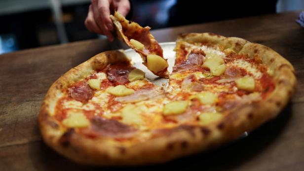 Eine Pizza mit Schinken und Ananas wird angeschnitten.