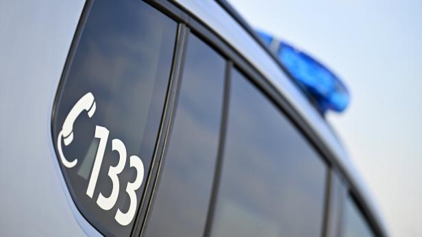 Auf der Autoscheibe ist in Weiß ein Telefonsymbol und die Nummer 133 zu sehen, im Hintergrund ein Blaulicht.