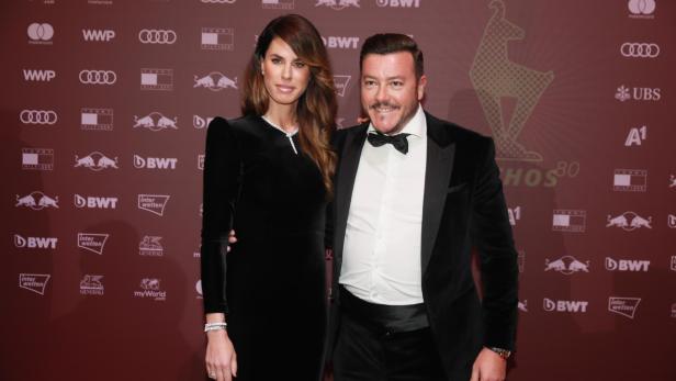 Ein Mann im Smoking und eine Frau im schwarzen Kleid posieren auf einem roten Teppich.