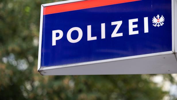 Blaues Schild mit weißer Aufschrift „Polizei“ und einem Adlerwappen, im Hintergrund unscharfe Bäume.