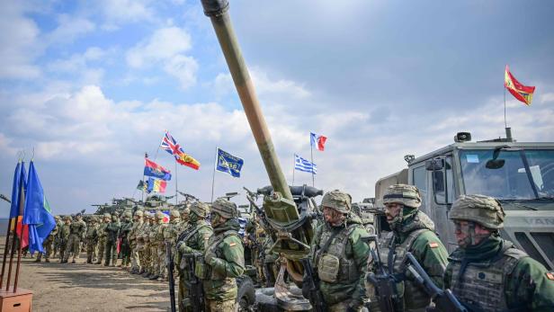 Soldaten stehen vor Militärfahrzeugen und Flaggen verschiedener Nationen.