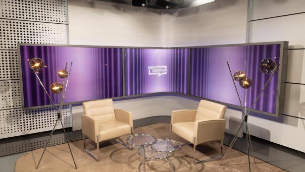 Ein Fernsehstudio mit zwei Sesseln und einem Tisch vor einem lila Bildschirm.