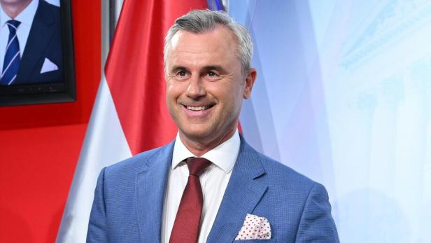 Ein lächelnder Mann im blauen Anzug mit roter Krawatte steht vor einer rot-weiß-roten Flagge.