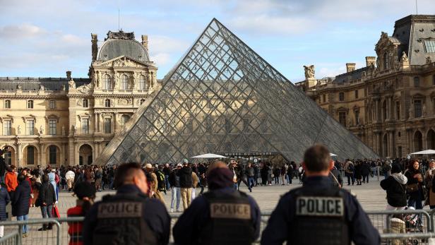Drei Polizisten stehen vor Absperrgittern, während viele Menschen vor der Glaspyramide des Louvre versammelt sind.