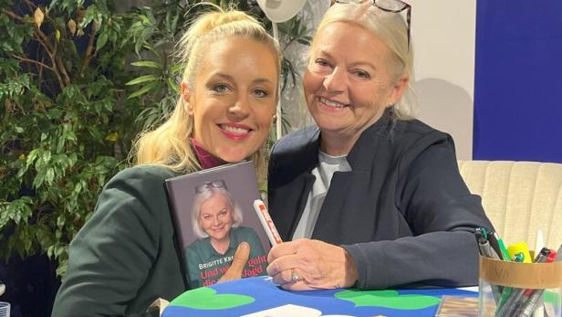 Zwei Frauen sitzen lächelnd zusammen, eine hält ein Buch mit dem Porträt der anderen, auf einem bunt gedeckten Tisch.