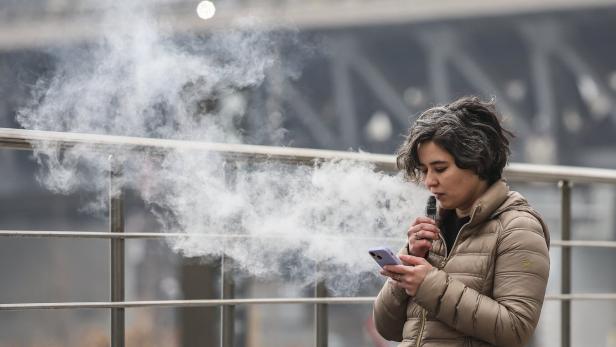 Eine Frau mit brauner Jacke benutzt eine E-Zigarette und schaut auf ein Smartphone, während viel Dampf entsteht.