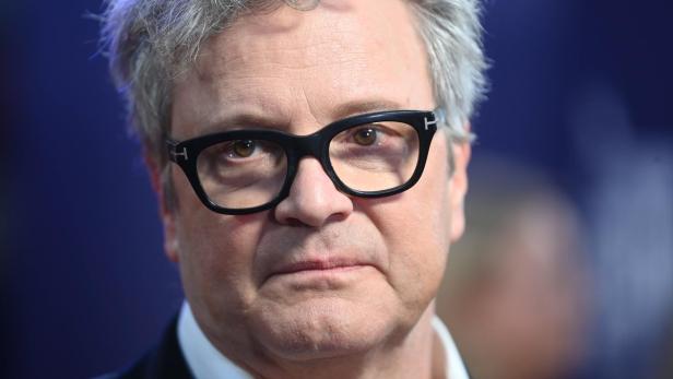 Colin Firth mit grauen Haaren und schwarzer Brille blickt in die Kamera.