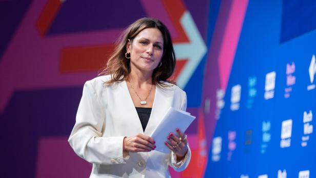 Shila Behjat mit weißem Blazer hält Papiere in der Hand und steht vor einer bunten Wand mit Logos und Schriftzügen.