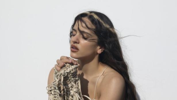 Rosalia mit langen dunklen Haaren trägt ein geflochtenes, cremefarbenes Kleid mit Fransen vor weißem Hintergrund.