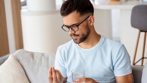 Ein Mann mit Brille hält ein Glas Wasser in der einen und eine gelbe Kapsel in der anderen Hand.