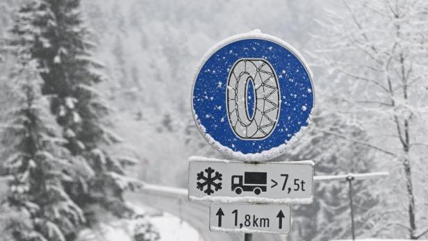 Ein blaues Schneeketten-Verkehrsschild mit Zusatztafeln für Lkw über 7,5 Tonnen steht in verschneiter Winterlandschaft.