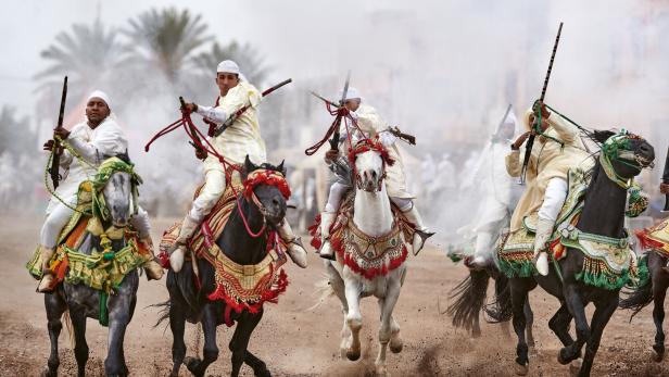 Mehrere Reiter in traditioneller Kleidung reiten auf geschmückten Pferden und halten Gewehre in die Luft, Staub wirbelt auf.