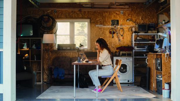 Person sitzt an einem Tisch in einer Werkstatt mit Holzverkleidung, Laptop, Waschmaschine und Werkzeug an der Wand.