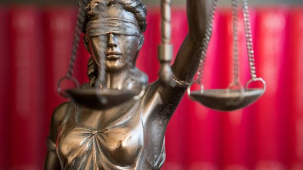 Eine Justitia-Statue mit Augenbinde und Waage steht vor einem roten Hintergrund.