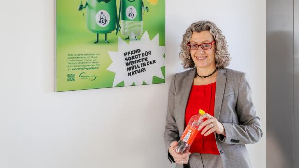 Eine Frau mit grauen Locken und rotem Oberteil hält eine Pfandflasche vor einem Plakat zum Thema Recycling.