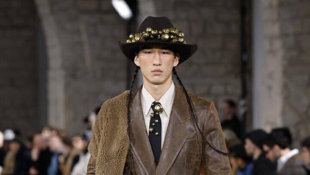 Ein Model mit Hut und Zöpfen präsentiert braune Lederkleidung auf einem Laufsteg.